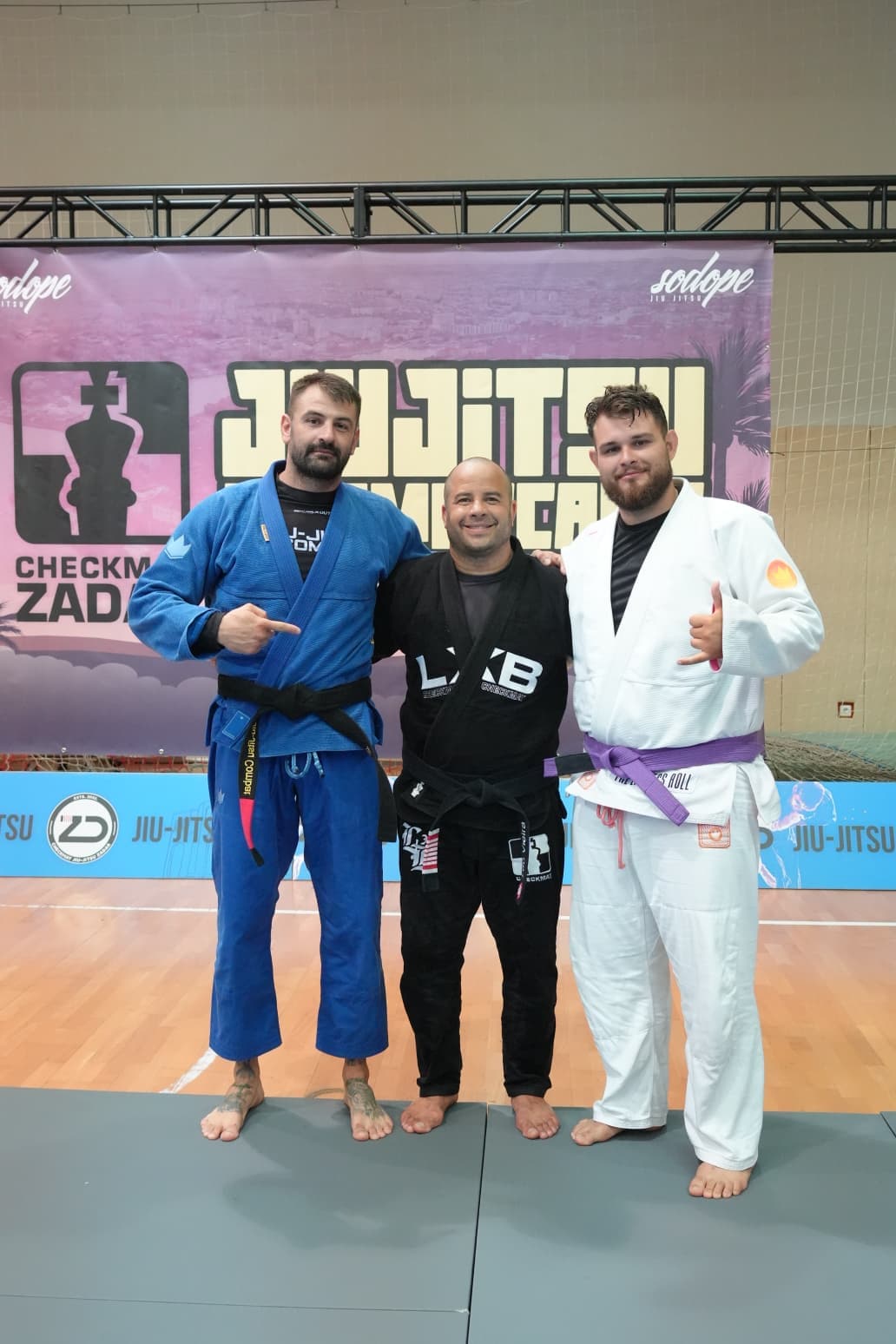 Trei sportivi în kimono la o competiție, în fața bannerului de turneu
